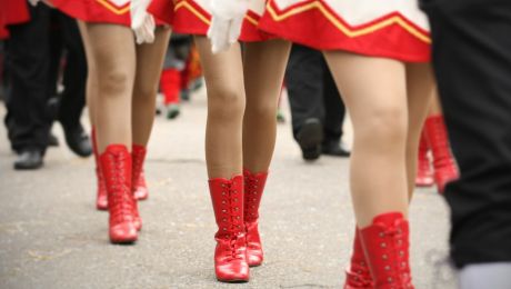Drei Frauenbeine in Gardeuniformen laufen hintereinander auf einer Straße. Sie tragen rote Stiefel.  