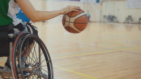 Eine Person in einem Sportrullstohl dribbelt mit einem Basketball in einer Sporthalle.