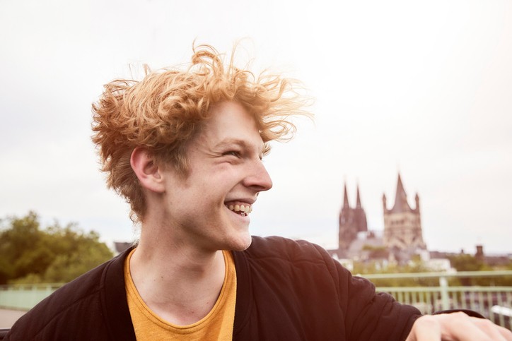 Junger Mann mit roten Haaren, links im Bild lacht und blick nach rechts. Kölner Dom verschwommen im Hintergrund.