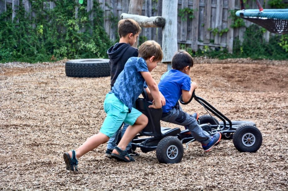 Ein Junge fährt Kettcar auf einem weitläufigen Spielplatz, während ihn zwei weitere Jungs anschieben.