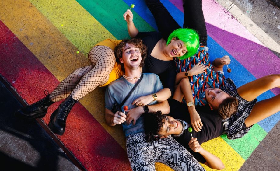 Vier Personen liegen aneinander gelehnt und lachend auf einer Progress Pride Flagge, die auf die Straße gemalt wurde.